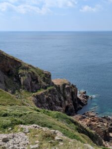 Vue en plongée depuis une falaise couverte de lande et de fleurs blanches sur un chaos de rochers sombres aux strates verticales plongeant dans une mer bleu-vert, avec un sentier visible sur la crête et l'océan Atlantique s'étendant à l'horizon, sous un ciel bleu légèrement voilé.