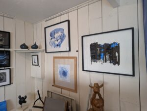 Intérieur d'une galerie d'art aux murs en lambris blanc, présentant plusieurs œuvres abstraites encadrées à dominante bleu-noir, des sculptures en céramique sombre sur une étagère et des figurines en bois, dans un espace intimiste et lumineux.