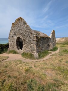 Ruines d'une petite chapelle romane en moellons de granite et de schiste, dont il ne reste que les murs gouttereaux et le pignon percé d'une baie en plein cintre, posées sur une lande herbeuse dominant la mer turquoise et les dunes de Carteret, sous un ciel bleu strié.