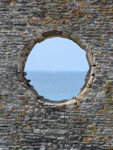 Vue à travers l'oculus circulaire en moellons de schiste et de granite couvert de lichen de la chapelle Saint-Germain, encadrant la Manche d'un bleu pâle avec un voilier blanc solitaire au centre, sous un ciel légèrement voilé.