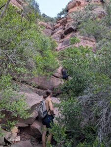 Deux randonneurs progressant en escalade dans un étroit couloir rocheux aux parois de grès rouge, entouré d'une végétation méditerranéenne dense de lauriers-roses et d'arbustes, l'un en bas s'apprêtant à grimper, l'autre déjà engagé plus haut sur la paroi.