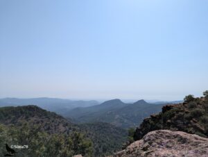 Vue panoramique depuis le sommet de l'El Garbí sur les reliefs boisés de la Serra Calderona, avec plusieurs sommets arrondis couverts de pins s'étirant vers une plaine lointaine estompée par la brume de chaleur estivale, sous un ciel bleu pâle et uniforme.