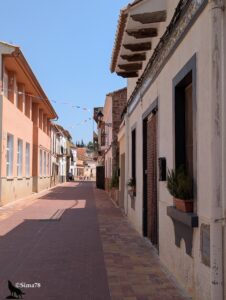 Rue principale pavée de briques rouges du village de Segart, bordée de maisons aux façades crépies blanches et ocre avec des toits de tuiles romanes sur consoles en bois, décorée de guirlandes de fanions colorés traversant la chaussée, déserte sous un soleil de plein midi, sous un ciel bleu vif.