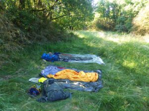 Deux couchages de bivouac installés côte à côte dans une clairière herbeuse en lisière de forêt, composés de matelas de sol argentés, d'un sac de couchage orange et de couvertures de survie, avec sacs à dos et équipement dispersés au premier plan, en pleine lumière de juin.
