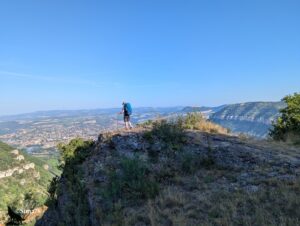 Randonneur de dos, coiffé d'un chapeau de randonné et portant un grand sac à dos bleu avec des bâtons de marche, debout sur un affleurement rocheux calcaire en surplomb dominant la vallée du Tarn, avec Millau et ses environs visibles en contrebas et les falaises des causses environnants à l'horizon, sous un ciel bleu parfaitement dégagé.