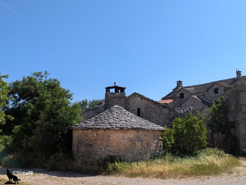 Édifice cylindrique en moellons calcaires coiffé d'un toit conique en lauzes grises surmonté d'un lanternon en pierre, apparemment attenant à une ferme caussenarde aux toits de lauzes, entourés de végétation, sous un ciel bleu vif.