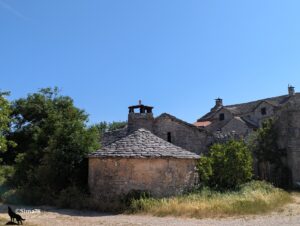 Édifice cylindrique en moellons calcaires coiffé d'un toit conique en lauzes grises surmonté d'un lanternon en pierre, apparemment attenant à une ferme caussenarde aux toits de lauzes, entourés de végétation, sous un ciel bleu vif.