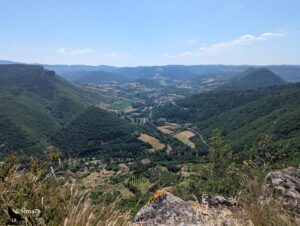 Vue panoramique en plongée depuis le rebord du causse du Larzac sur la vallée verdoyante de la Dourbie encaissée entre des versants boisés, avec le bourg de Nant visible au centre dans le fond de vallée agricole, des reliefs tabulaires à l'horizon, sous un ciel bleu de juin.