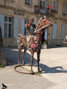 Sculpture grandeur nature d'un chevalier en armure monté sur un cheval, entièrement assemblée à partir de pièces métalliques recyclées et d'objets de récupération oxydés, installée sur la place devant la mairie de Nant, bâtiment en pierre calcaire aux volets gris, sous un soleil estival.