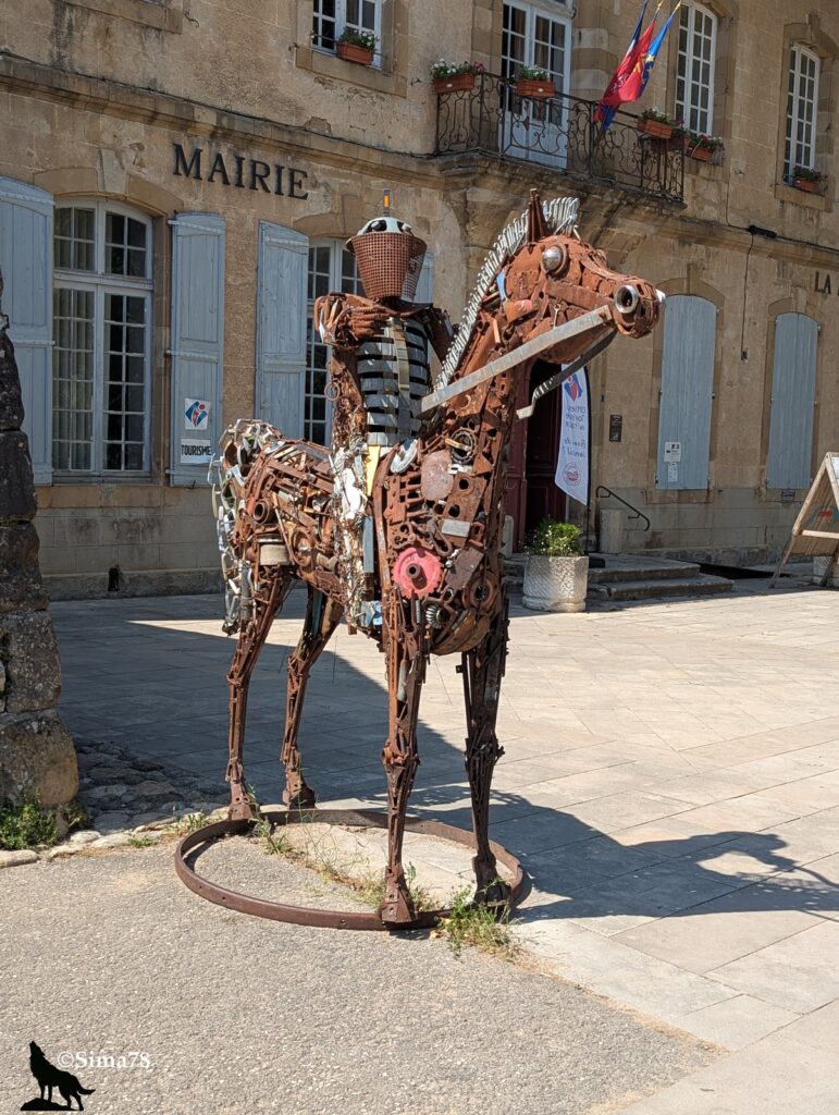 Sculpture grandeur nature d'un chevalier en armure monté sur un cheval, entièrement assemblée à partir de pièces métalliques recyclées et d'objets de récupération oxydés, installée sur la place devant la mairie de Nant, bâtiment en pierre calcaire aux volets gris, sous un soleil estival.