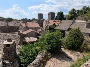 Vue en plongée depuis les remparts sur les toits de lauzes et de tuiles romanes du village médiéval de La Couvertoirade, avec ses deux tours carrées crénelées dominant l'ensemble des maisons en pierre calcaire, des arbres et une terrasse de café ombragée, sous un ciel bleu à cumulus.
