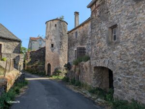 Ruelle goudronnée étroite d'un hameau caussenard bordée de maisons en moellons calcaires, avec une tour cylindrique en pierre percée d'une porte en arc et d'une archère, partiellement en ruine, sous un ciel bleu vif de juin.