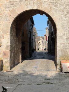 Vue depuis l'extérieur à travers une large porte en arc brisé en pierre calcaire donnant sur la rue principale pavée de La Cavalerie, village médiéval fortifié, avec des maisons en pierre, des lanternes et une boulangerie visibles dans la ruelle déserte, sous un ciel bleu matinal.