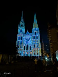 Vue nocturne de la façade occidentale de la cathédrale Notre-Dame de Chartres entièrement couverte d'un mapping lumineux aux teintes bleu turquoise, or et rose, illuminant ses deux flèches asymétriques, sa rosace et ses trois portails sculptés, devant un ciel noir et un public de spectateurs silhouettés au premier plan.