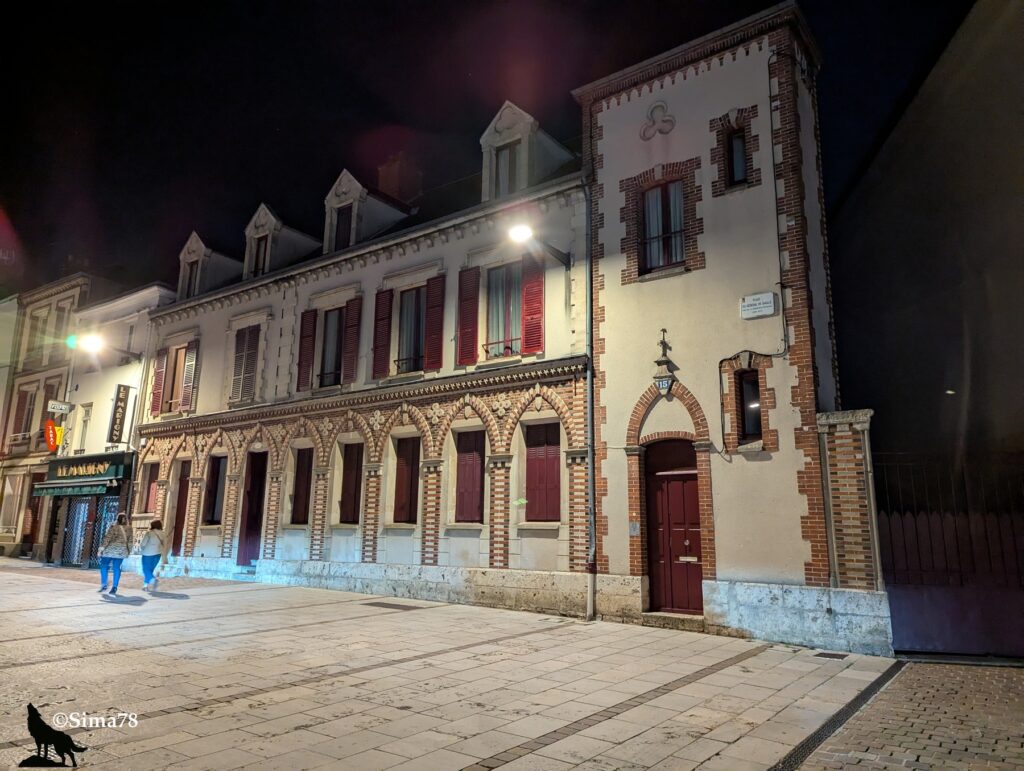 Bâtiment historique en brique et pierre, éclairé la nuit, avec des arcades et des fenêtres à volets rouges, situé Place du Général de Gaulle à Chartres.