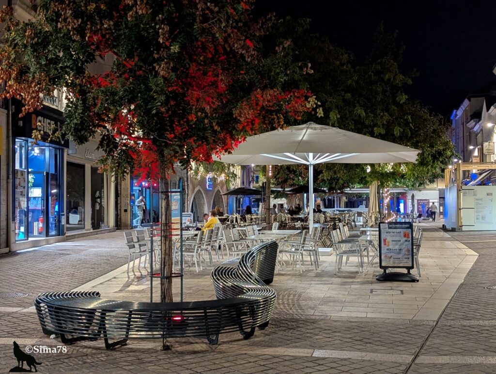 Terrasse de café éclairée la nuit, avec des tables, des chaises et des parasols blancs, entourée d’arbres illuminés en rouge, dans une rue piétonne pavée à Chartres.
