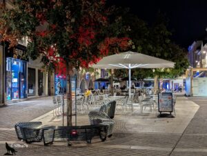 Terrasse de café éclairée la nuit, avec des tables, des chaises et des parasols blancs, entourée d’arbres illuminés en rouge, dans une rue piétonne pavée à Chartres.