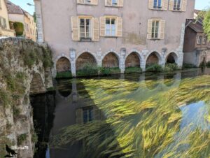 Ancien lavoir en pierre avec des arches, bordé par une maison à volets beiges, reflété dans l’eau de l’Eure, avec des herbes aquatiques flottant à la surface.