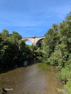 Pont en pierre à deux arches au-dessus de l’Eure, entouré d’une végétation dense et de berges herbeuses, sous un ciel bleu clair.