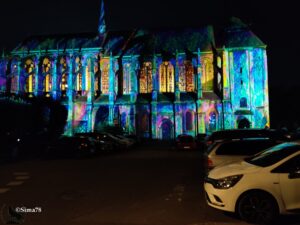 Façade de l’Église Saint-Pierre de Chartres illuminée par des projections colorées (bleu, violet, turquoise), avec ses vitraux et ses arcs mis en valeur, de nuit, et des voitures garées en contrebas.