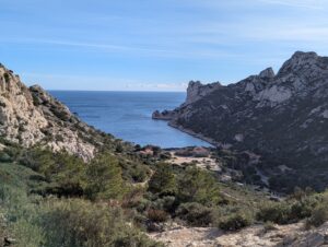 Vue plongeante depuis un sentier calcaire sur la calanque de Sormiou, encadrée par deux falaises blanches aux parois déchiquetées couvertes de pinèdes, avec le fond de calanque et ses quelques habitations visibles en contrebas et la Méditerranée bleue s'ouvrant vers l'horizon, sous un ciel bleu strié de traînées légères.