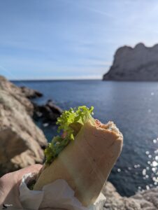 Main tenant un sandwich garni de salade verte en gros plan, avec en arrière-plan flou la Méditerranée scintillante, des rochers calcaires et la falaise blanche de la calanque de Sormiou sous un ciel bleu hivernal.