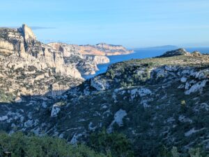 Vue panoramique depuis les hauteurs du massif des Calanques sur une succession de falaises calcaires plongeant vers la Méditerranée bleue, avec le Cap Canaille aux teintes ocre et dorées visible au loin, une île silhouettée à l'horizon, et un vallon boisé de pins en contrebas, dans la lumière dorée de fin d'après-midi de janvier.