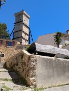 Tour médiévale en pierre de Saint-Martin-de-Brômes, consolidée par une structure métallique, vue depuis une ruelle en escalier avec murets en pierre sèche, sous un ciel bleu.