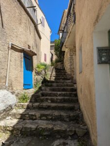 Ruelle en escalier de pierre dans le village de Saint-Martin-de-Brômes, encadrée de maisons provençales, avec une porte peinte en bleu vif sur la gauche, sous un ciel bleu.