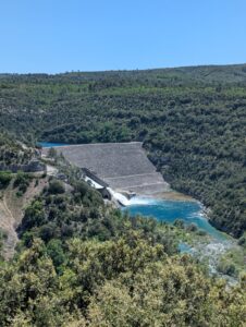 Vue plongeante depuis la crête du Lourmant sur le barrage d'Esparron-de-Verdon, avec son parement en enrochements gris, sa vanne ouverte déversant un puissant jet d'eau blanc dans le Verdon aux eaux turquoise, encadré de collines boisées.