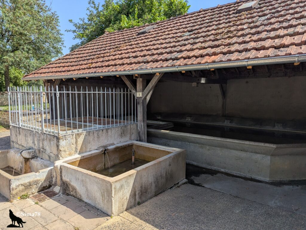 Photographie d'un lavoir couvert traditionnel en pierre, avec toit de tuiles et bassins alimentés par des fontaines, situé sur une place de la Fontaine.