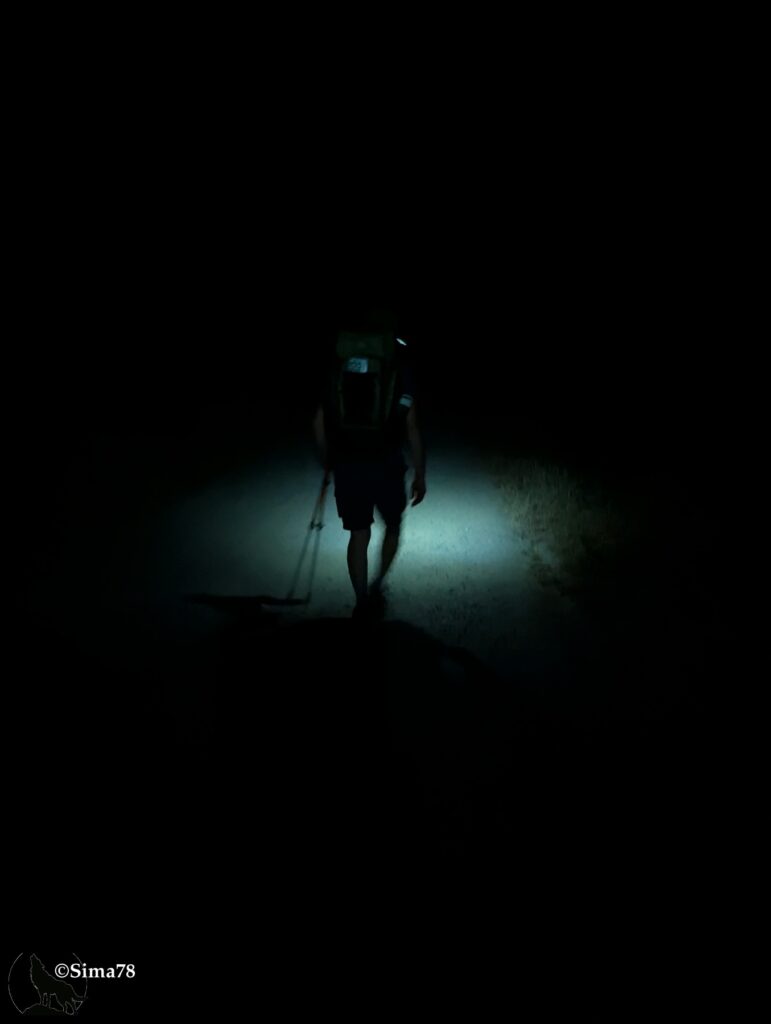 Photographie de nuit d'un randonneur de dos, sac à dos et bâtons de marche, éclairé par une lampe frontale sur un chemin plongé dans l'obscurité.