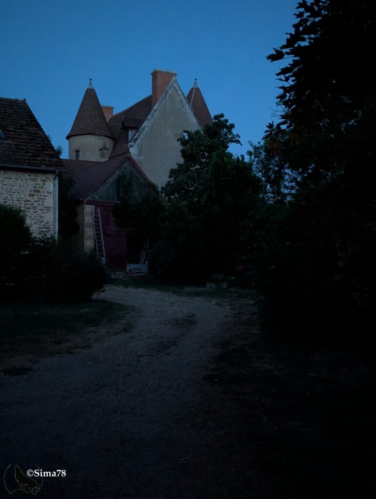 Photographie de nuit d'un chemin de gravier traversant un hameau, avec une tourelle et un pignon de bâtiment ancien se détachant sur un ciel bleu nuit encore sombre.