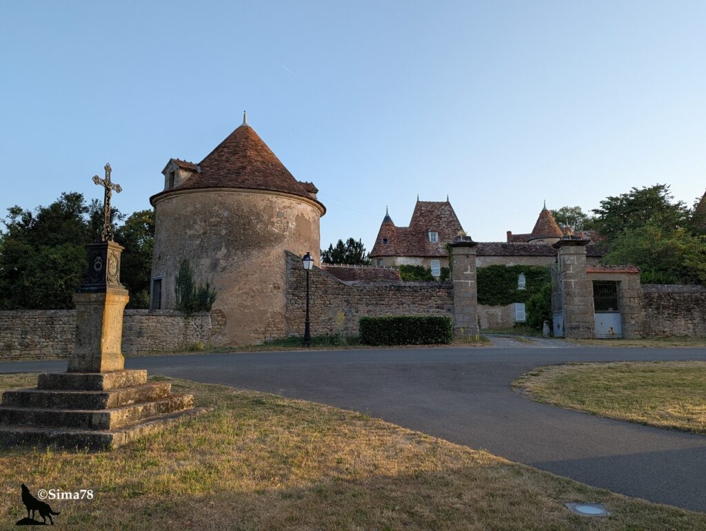 Photographie du château de La Pouzerie au petit matin, avec un pigeonnier rond à toit conique en tuiles, une croix de chemin en pierre, un mur d'enceinte et un portail d'entrée, sous un ciel bleu dégagé.