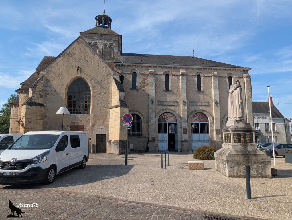 Photographie d'un ancien édifice religieux roman reconverti en musée, avec clocher à coupole, arcades en pierre et enseignes indiquant « Musée des Vieux Métiers », un monument aux morts au premier plan, sous un ciel bleu dégagé.