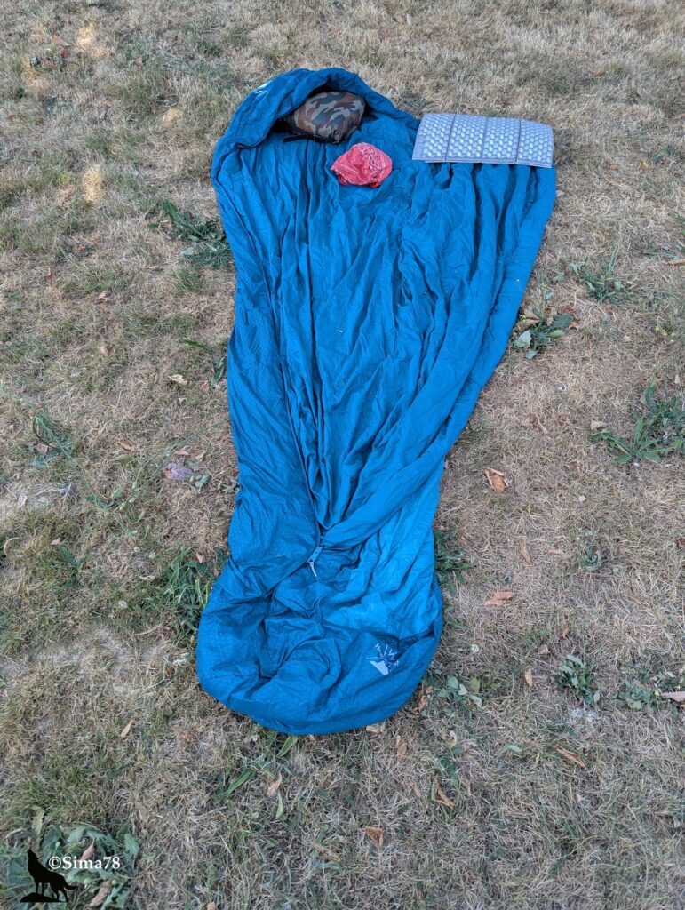 Photographie d'un sac de couchage bleu déplié à même l'herbe d'un camping, servant de matelas, avec un bandana rouge, un tapis de sol et un vêtement posés dessus.