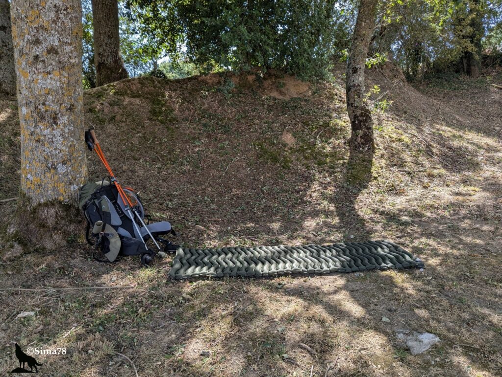 Photographie d'un sac à dos, de bâtons de marche et d'un matelas de sol gonflable installés à l'ombre d'arbres, au pied d'un talus, dans une clairière forestière.