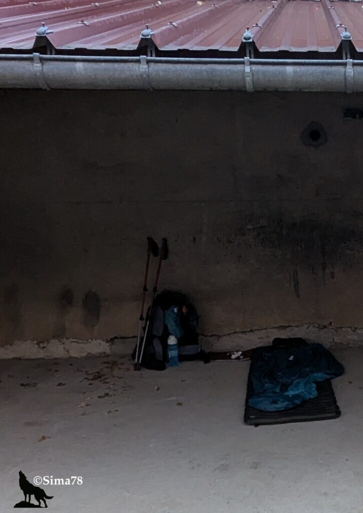 Photographie d'un sac à dos, de bâtons de marche et d'un sac de couchage installés au sol sous un abri en tôle, dans une lumière tamisée de petit matin.