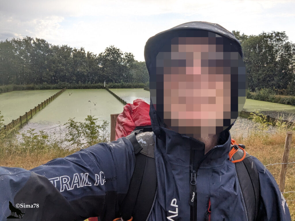 Photographie d'un randonneur en veste de pluie et capuche, sac à dos équipé d'une housse de protection rouge, devant un étang bordé de piquets de bois sous une pluie fine.