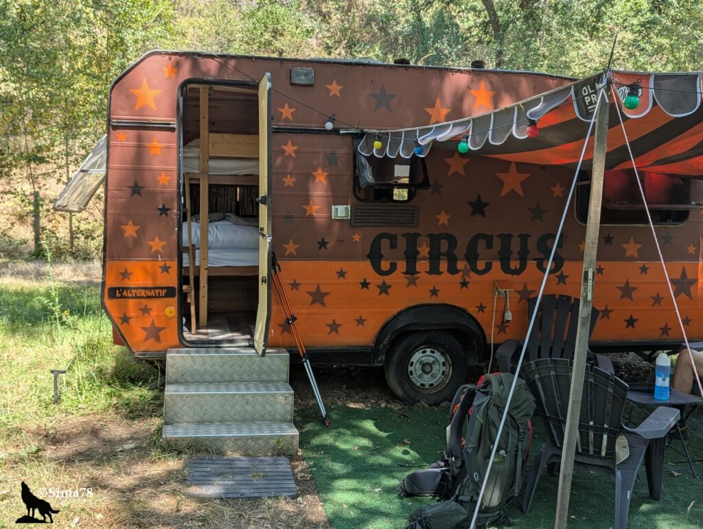 Photographie d'une caravane vintage décorée d'étoiles orange et noires et de l'inscription « Circus », aménagée en hébergement insolite, avec ses marches d'accès et une guirlande lumineuse, dans un cadre boisé.