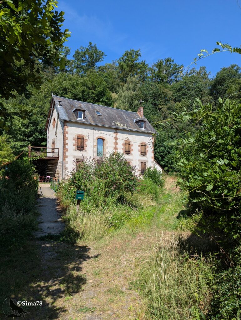 Photographie d'une ancienne maison de meunier en pierre et brique, avec toit d'ardoise et lucarnes, nichée au creux d'un vallon boisé et accessible par un chemin herbeux.