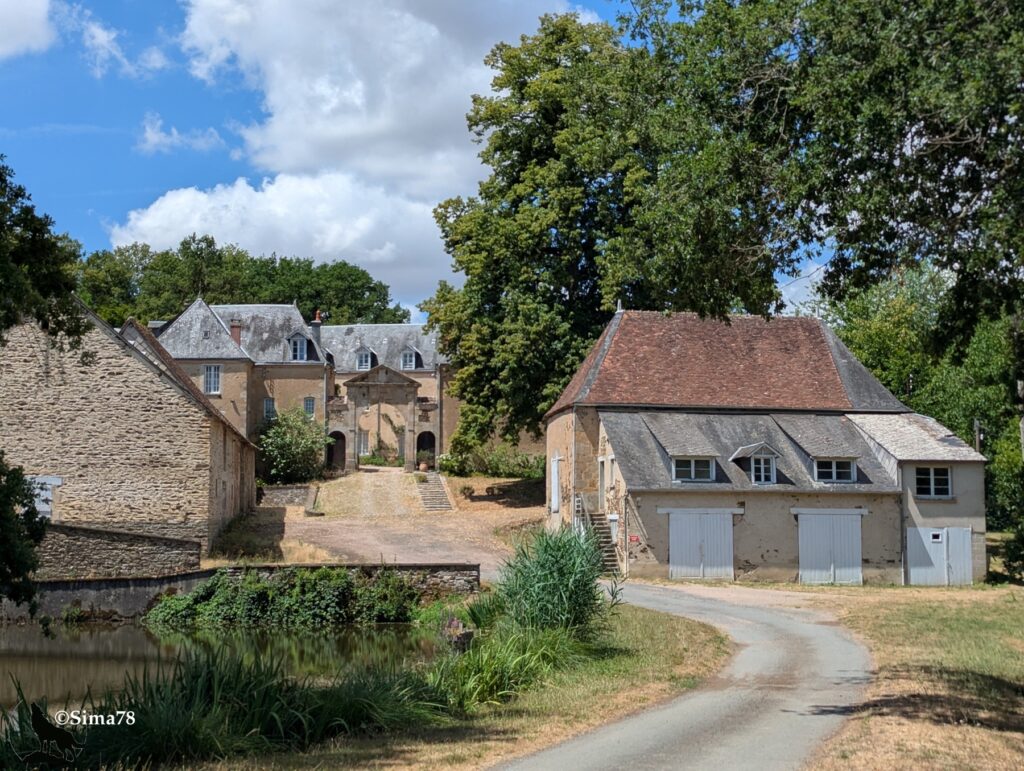 Photographie du château de Beaulieu et de ses dépendances en pierre, avec cour d'honneur, portail d'entrée et bâtiments agricoles au toit de tuiles, bordés d'un étang et d'arbres.