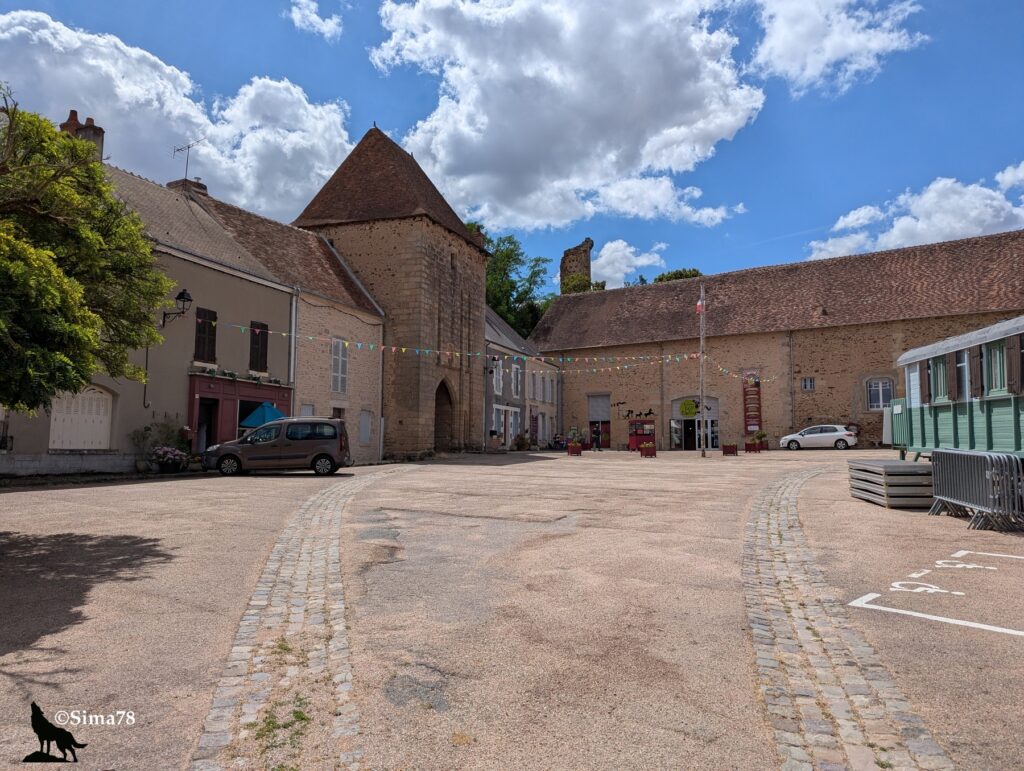 Photographie d'une place pavée bordée de bâtiments en pierre, dominée par une tour-porche fortifiée coiffée d'un toit conique en tuiles, décorée de guirlandes de fanions colorés, sous un ciel bleu parsemé de nuages.
