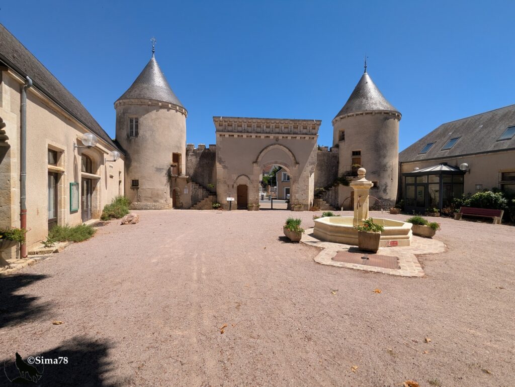 Photographie d'une cour intérieure ornée de deux tourelles coniques encadrant un porche voûté, avec une fontaine en pierre au premier plan, sous un ciel bleu dégagé.