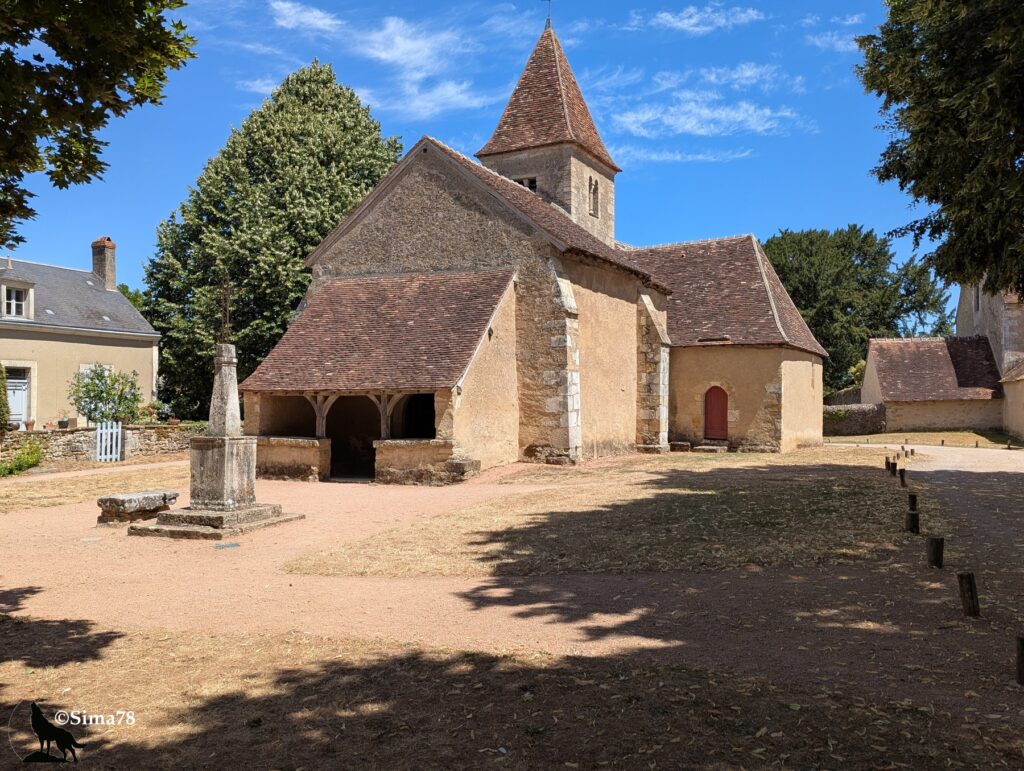 Photographie de l'église de Nohant, édifice en pierre au clocher pointu recouvert de tuiles, avec une croix de pierre ancienne au premier plan, sous un ciel bleu parsemé de nuages fins.