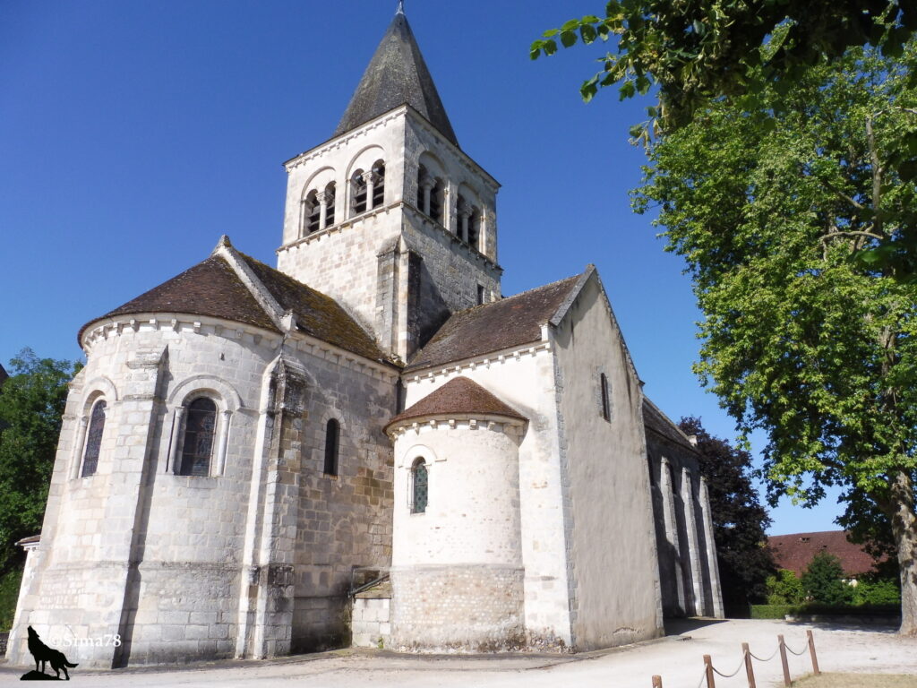 Photographie de l'église romane Notre-Dame de La Berthenoux, avec son clocher à flèche de pierre percé de baies géminées, son abside en cul-de-four et son chevet à contreforts, sous un ciel bleu dégagé.