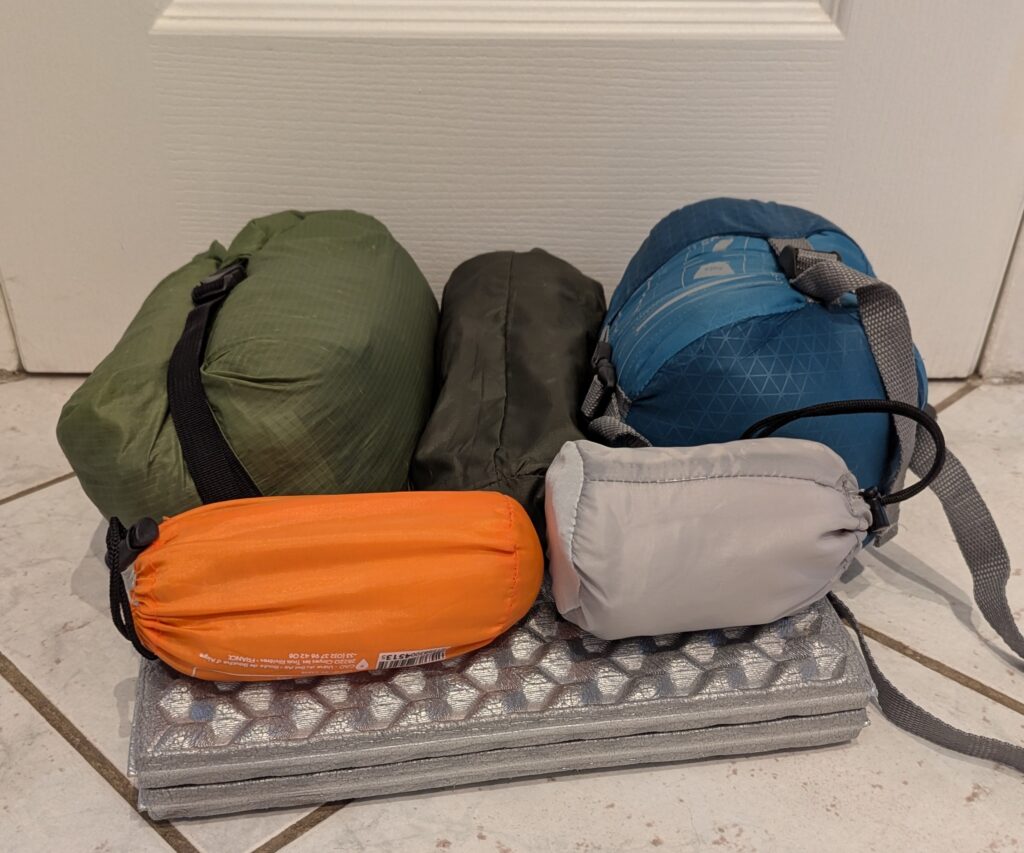 Photographie de matériel de randonnée compacté dans des housses, posé sur un coussin d'assise en mousse : au fond, de gauche à droite, la tente (housse verte), le matelas (housse kaki) et le sac de couchage (housse bleue) ; au premier plan, une housse orange contenant le film plastique isolant du sol et une housse grise contenant le sac à viande en soie.