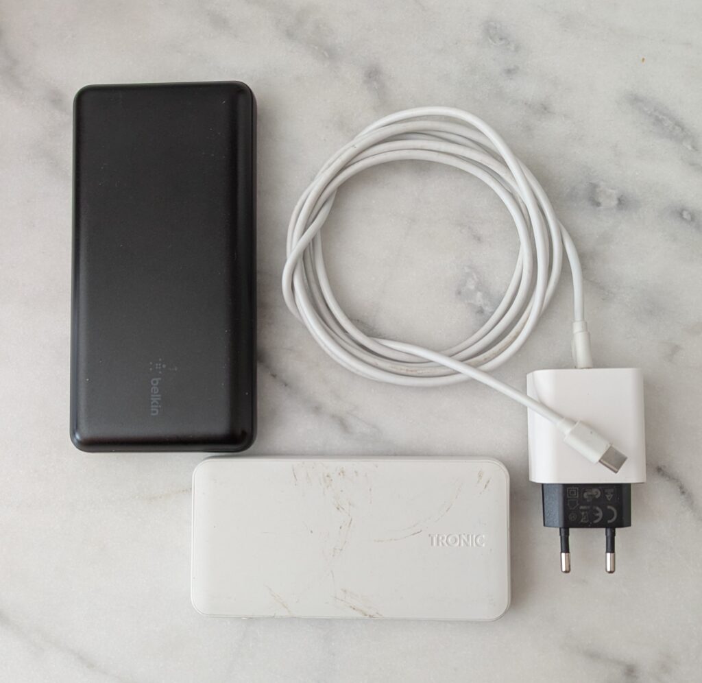 Photographie de deux batteries externes et d'un chargeur : à gauche, une batterie noire Belkin de format standard ; en bas, une batterie blanche TRONIC de format identique ; à droite, un chargeur secteur blanc avec câble USB-C enroulé et prise européenne.