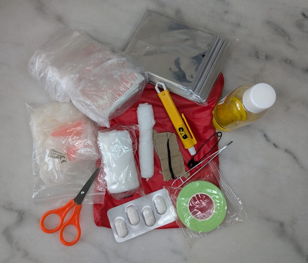 Photographie du contenu de la trousse de secours étalé sur une pochette rouge : une paire de ciseaux à bouts orange, une paire de gants en latex, un sachet contenant compresses et pansements classiques et spéciaux ampoules, une couverture de survie argentée, du sparadrap chirurgical, une plaquette de comprimés de paracétamol, un bandage neuf et un bandage entamé, une pince à tiques jaune, une pince à épiler, du fil noir et une aiguille, une épingle à nourrice, et un flacon d'huiles essentielles à base de jojoba.