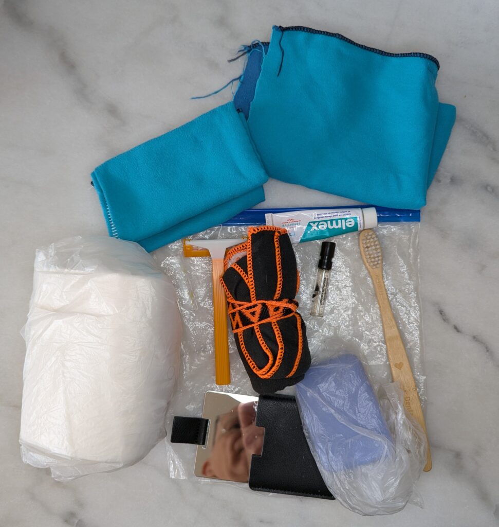 Photographie du contenu de la trousse de toilette étalé : un chiffon et un gant de toilette microfibre bleu turquoise, une serviette microfibre noire enroulée, un rouleau de papier toilette, un rasoir, un flacon échantillon de parfum, une brosse à dents en bois, un dentifrice Elmex, un miroir en acier poli et un savon.